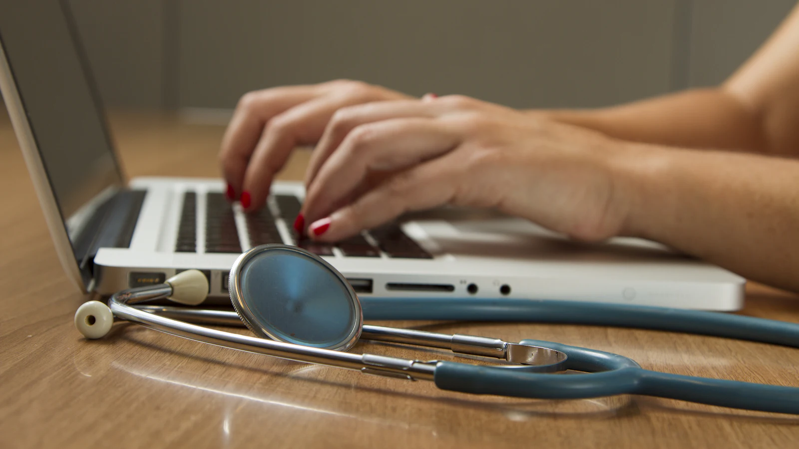 Hands typing on a laptop with a stethoscope on the desk
