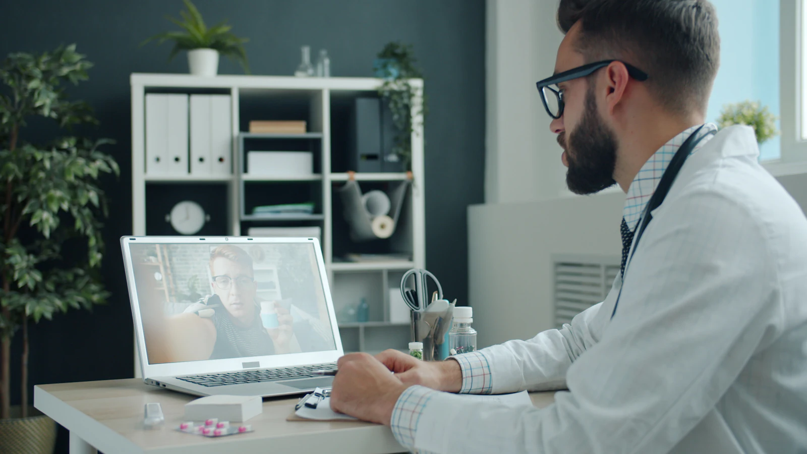 A doctor in an office consulting a patient by video call on a laptop